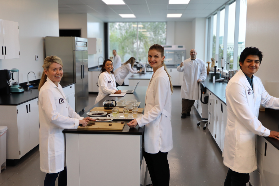 The M2 Center of Innovation is a food science lab and in this image you see the team standing at the tables with various scientific equipment and samples including coffee.
